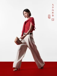 Model wearing a red top and white trousers holding a handbag against a white background with a red floor stripe.