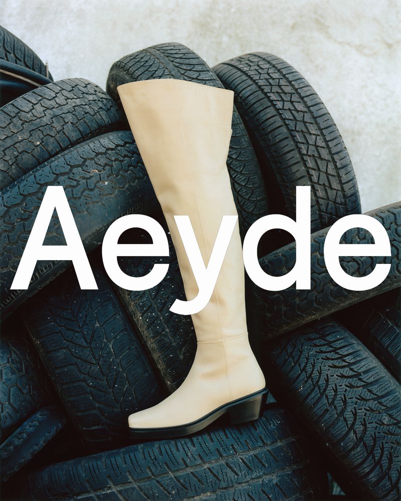 Beige over-the-knee boot displayed on a stack of black tires against a grey concrete backdrop, fashion-forward footwear.