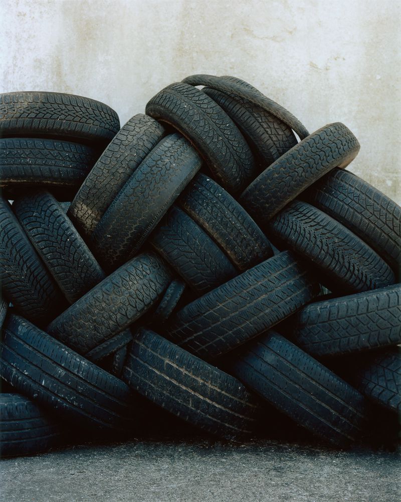 Stack of black rubber car tires arranged in a woven pattern against a neutral concrete background, creating a textured wall.