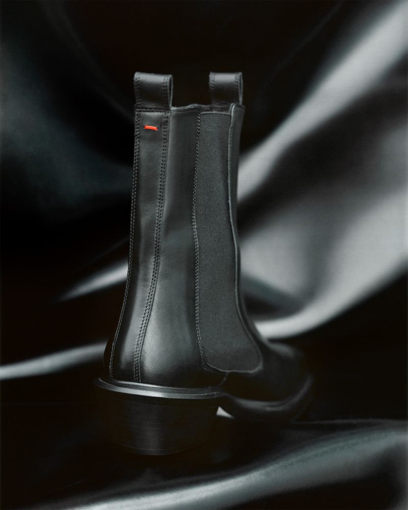 Studio shot of a black leather Chelsea boot with elastic side panels and a small red detail, set against a dark draped fabric backdrop.