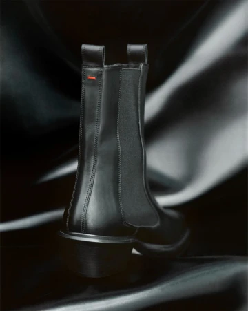 Studio shot of a black leather Chelsea boot with elastic side panels and a small red detail, set against a dark draped fabric backdrop.