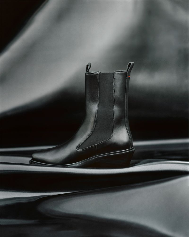 Studio shot of a black leather Chelsea boot with a wedge heel against a rippled, reflective gray and black background.