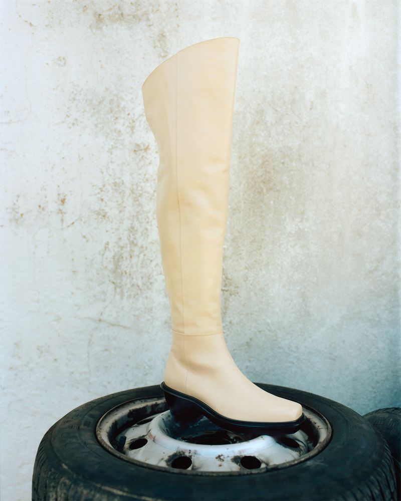 A single beige over-the-knee boot with a chunky black heel is featured against a neutral wall and sits on a tire.