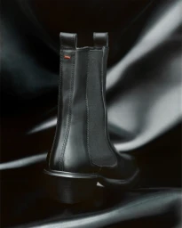 Black leather ankle boot with elastic side panels standing on a glossy dark draped fabric background.