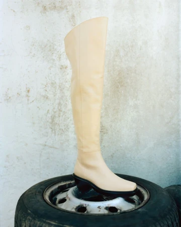 Cream over-the-knee leather boot resting upright on a black car tire against a textured grey wall.