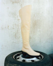 Cream over-the-knee leather boot resting upright on a black car tire against a textured grey wall.