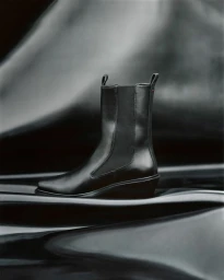 A black leather Chelsea boot from the Aeyde Biker Capsule collection resting on a dark draped metallic surface.