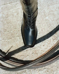 Top-down view of a black lace-up leather boot standing on a concrete ground with a coiled leather strap in the foreground