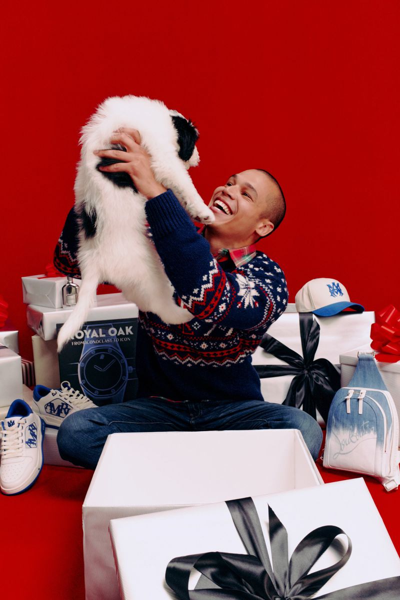 A man is holding a fluffy puppy in front of a bright red background, surrounded by white gift boxes, clothing and accessories.