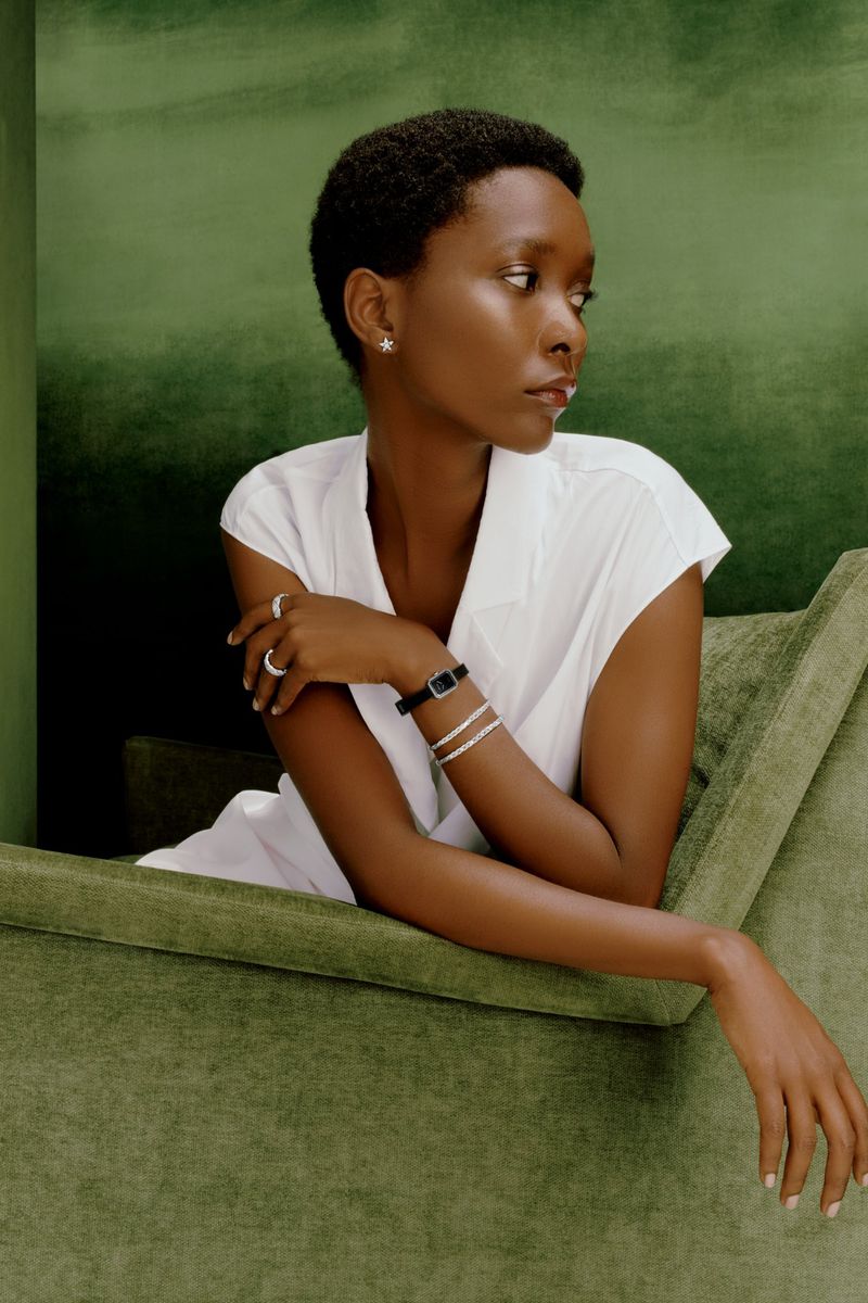 Elegant African-American woman in a white sleeveless top, with jewelry, seated on a green velvet chair against a green backdrop.