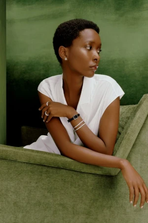 Elegant African-American woman in a white sleeveless top, with jewelry, seated on a green velvet chair against a green backdrop.