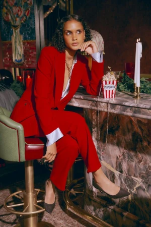 Elegant woman in a red suit poses in a bar setting with a popcorn bucket, seated on a high stool, creating a stylish, sophisticated image.