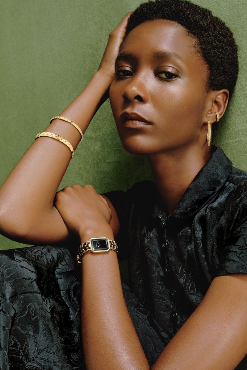Elegant portrait of a Black woman wearing jewelry and a watch against an olive green background in soft, diffused studio lighting.