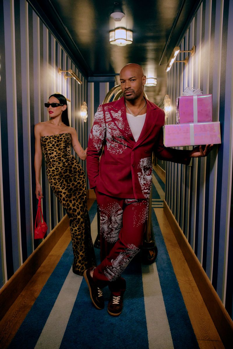 Two fashion models pose in a striped hallway. A male model holds two pink gift-wrapped boxes. The female model wears sunglasses.