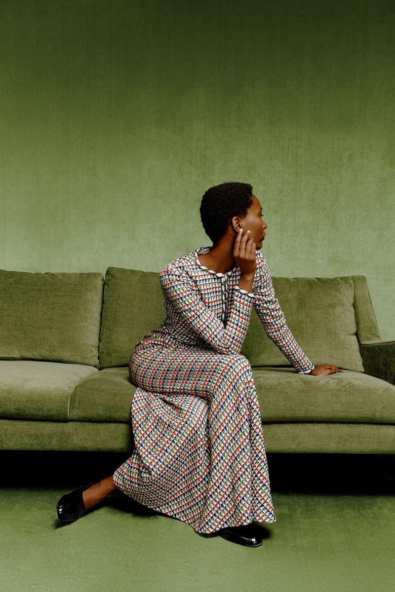 Adult woman with short, dark hair poses on a green velvet couch in a long-sleeved, patterned dress and black shoes against a green backdrop.