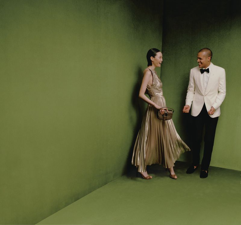 A stylish couple in formal wear stands in a green corner setting; the woman in a flowing dress with a decorative handbag, the man in a white tuxedo.