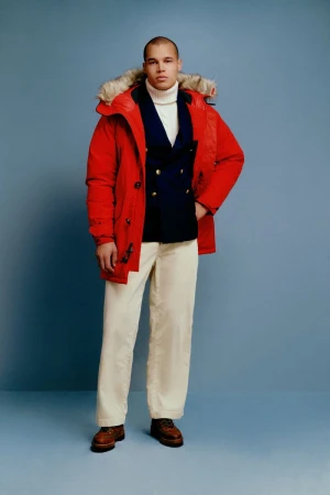 Man in red parka, navy blazer, and white pants strikes a confident pose against a light blue backdrop in a fashion shot.