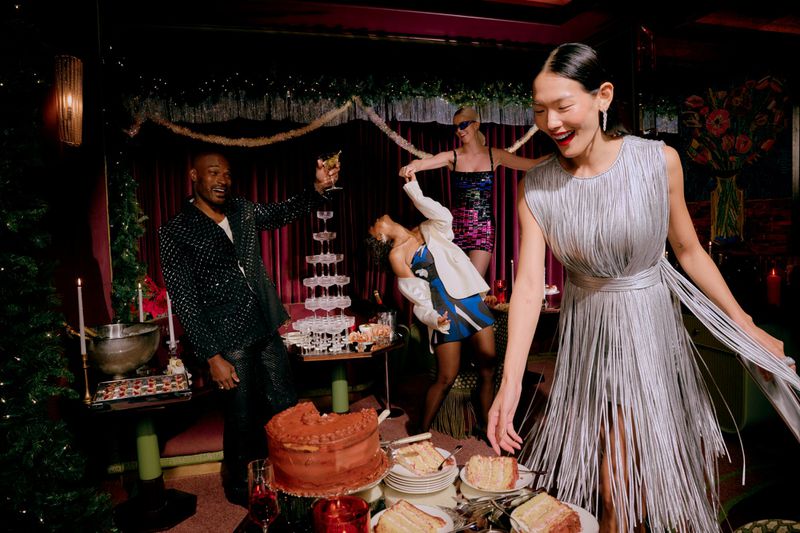 An Asian woman in a silver dress smiles at a party with other partygoers, a champagne tower, cake, and festive decorations.