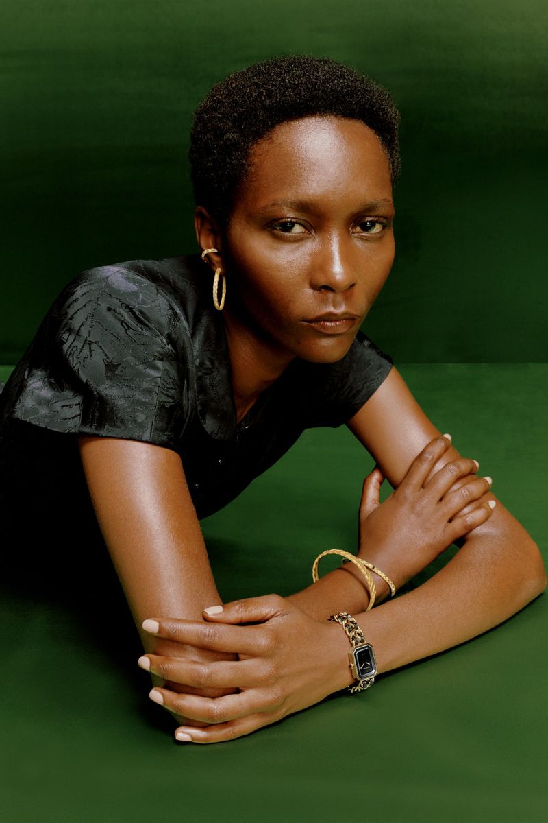 Woman with dark skin and short hair, wearing a black top, gold hoop earrings, a watch, and a bracelet, poses against a green background.