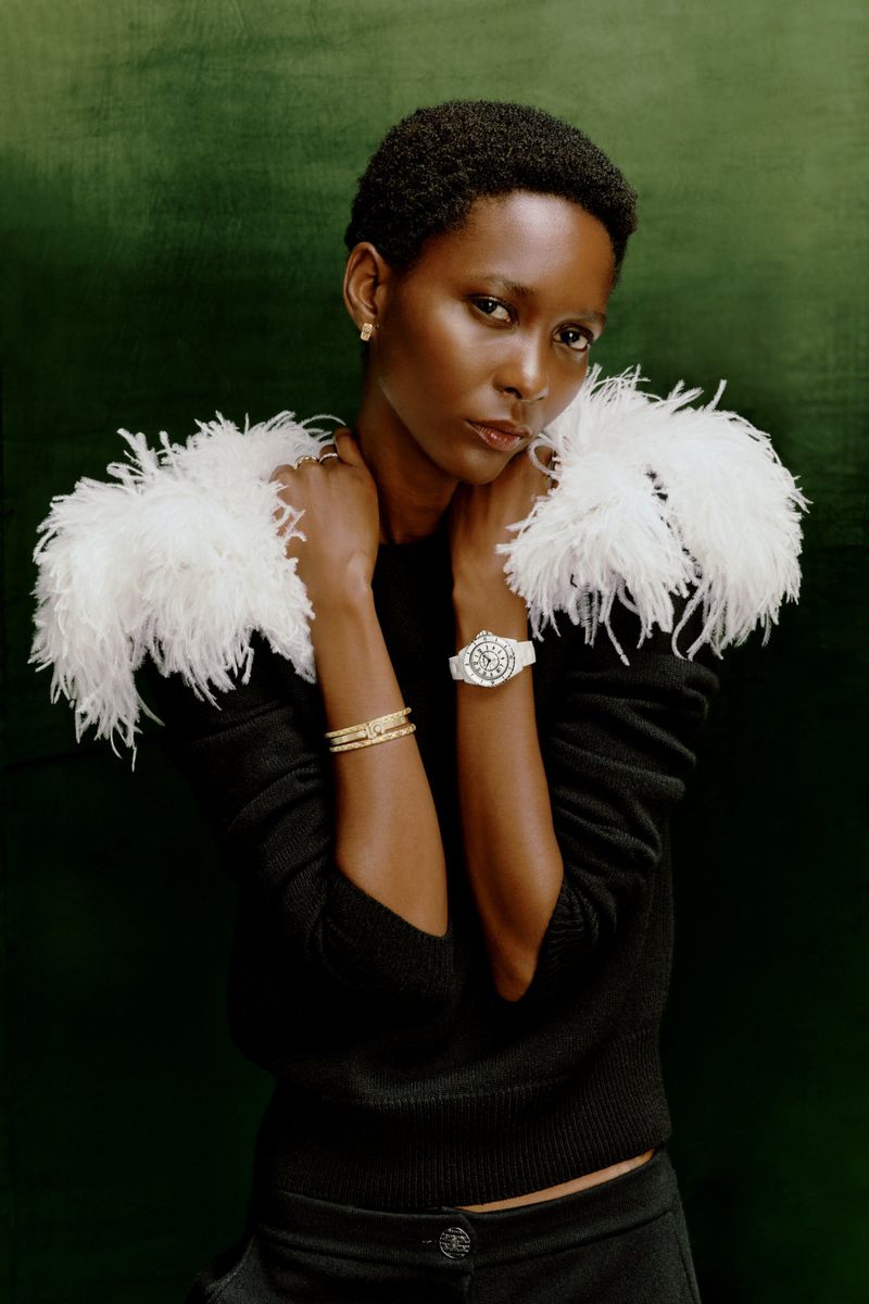Close-up of a stylish African-American woman wearing a black top, white feather boa, jewelry, and a white watch, set against a green backdrop.