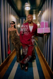 Tyson Beckford in a burgundy patterned suit holding gift boxes in a hotel hallway with a woman in a leopard dress