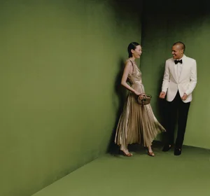 A woman in a gold pleated gown and a man in a white tuxedo jacket standing against a dark green studio wall.