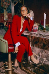 A woman in a red pantsuit sits on a barstool beside a marble bar with candles and popcorn for Saks Holiday 24.