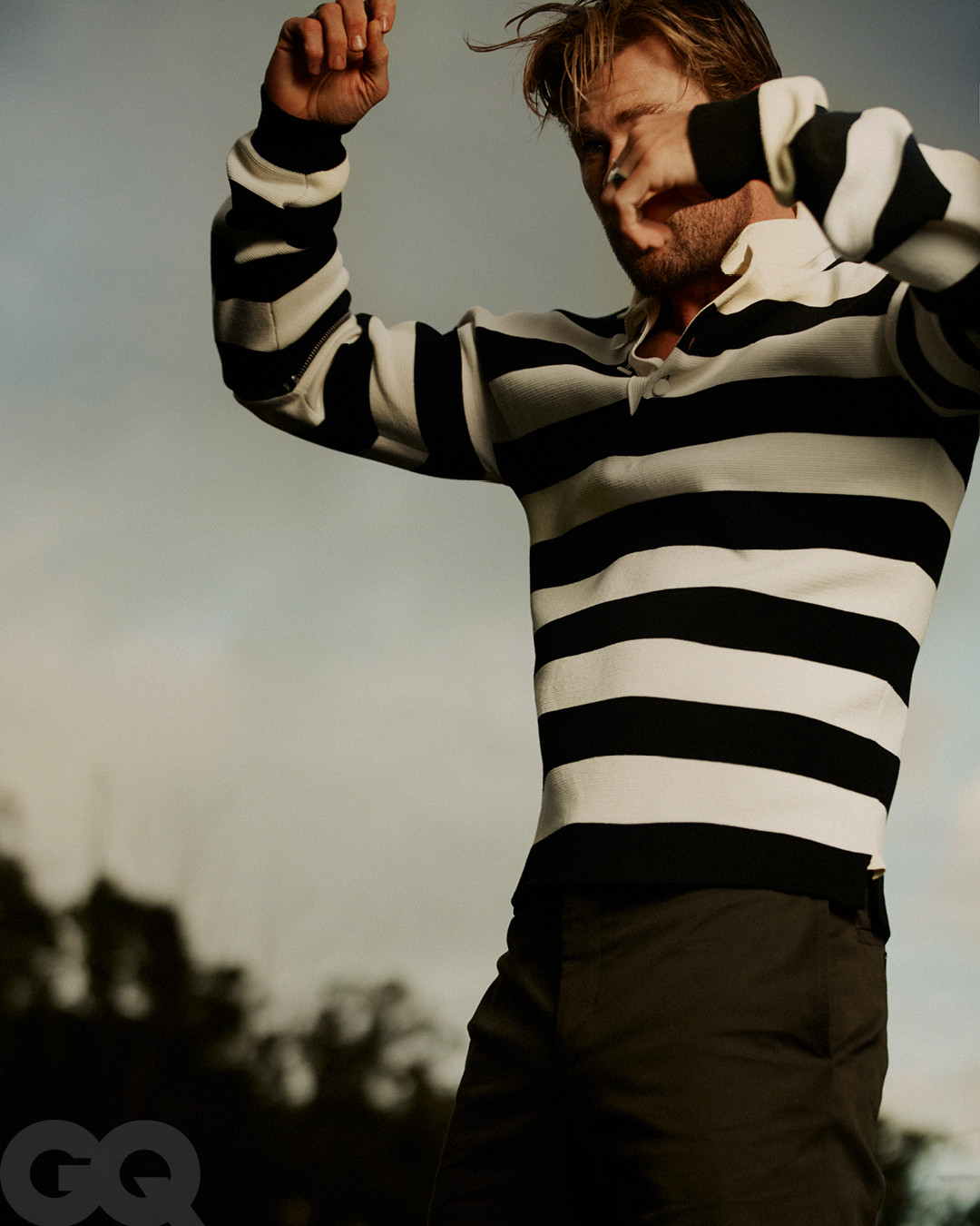 Portrait of a blonde adult man in a striped shirt with arms raised against a sky background, captured at eye-level.
