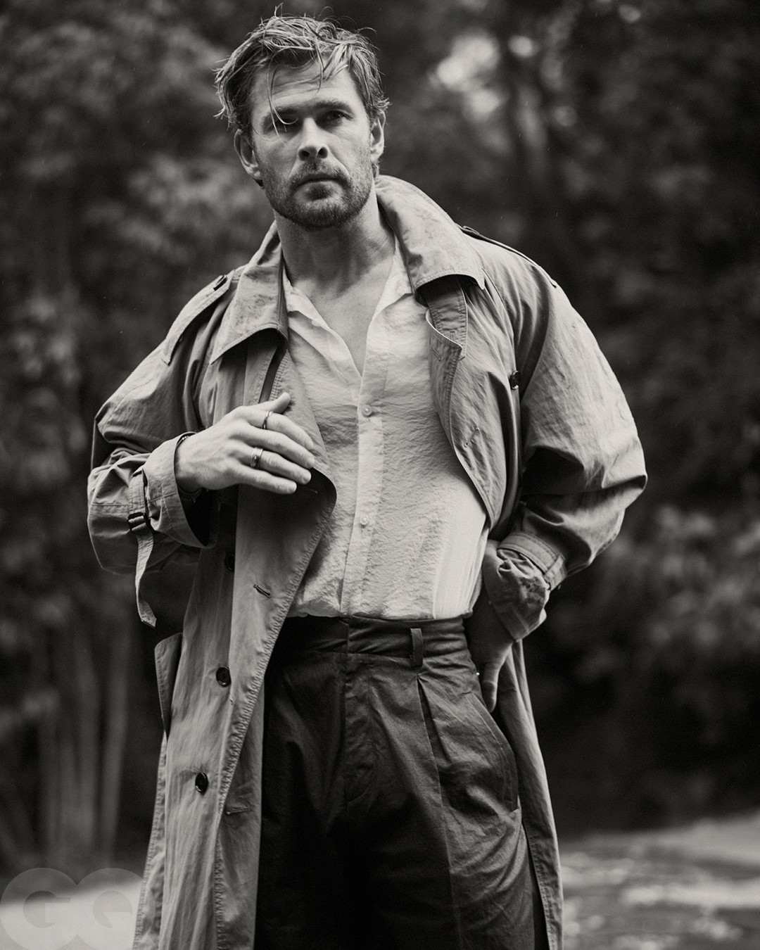 Monochrome portrait of male fashion model Chris Hemsworth in an open dress shirt, overcoat, and dress pants posing outdoors among trees.