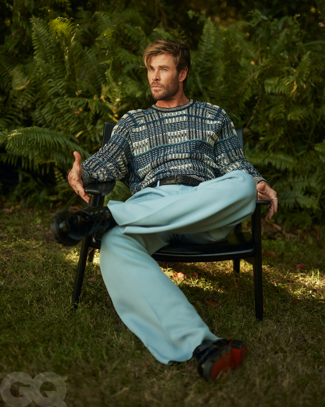 Chris Hemsworth sits in a chair outdoors, wearing a patterned sweater and light blue pants, on a green lawn with trees in the background.