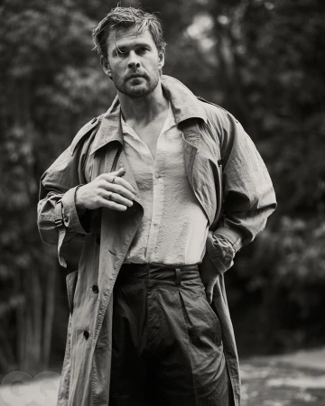 Monochrome portrait of Chris Hemsworth wearing an unbuttoned shirt and trench coat outdoors against a blurred woodland background