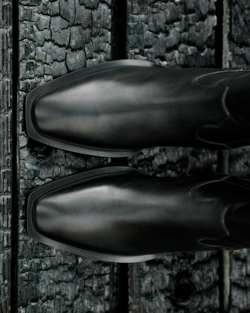 Close-up of two black leather western boots with square toes resting on a rough, dark wooden surface. Fashionable footwear.