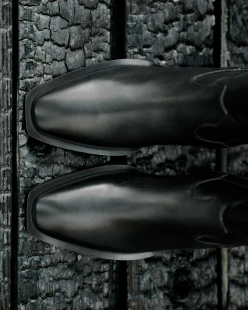 Close-up of two black leather western boots with square toes resting on a rough, dark wooden surface. Fashionable footwear.