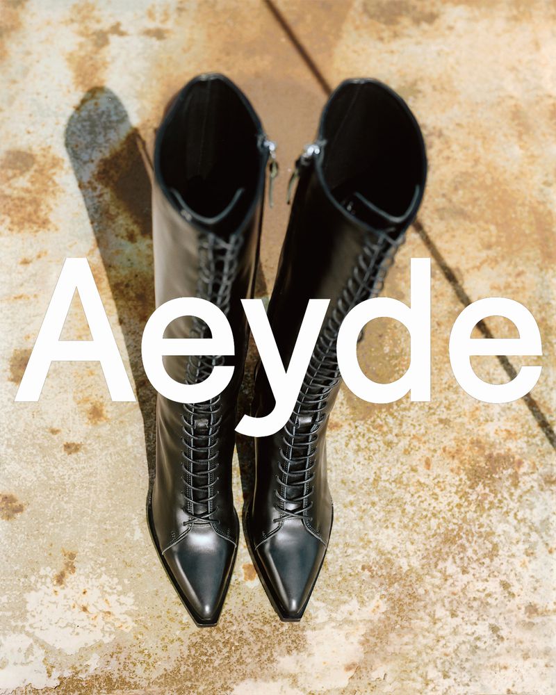 A pair of black, patent-leather Aeyde lace-up boots are showcased indoors with soft natural light and a minimalist aesthetic.