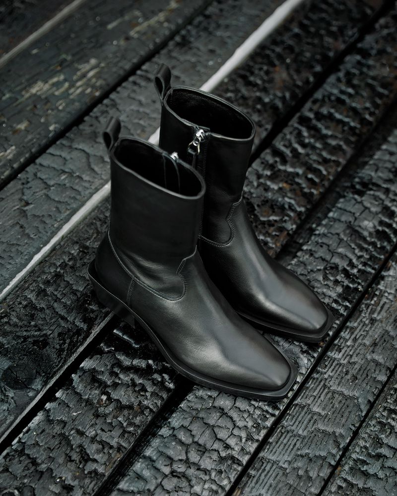 Pair of black leather ankle boots with a zipper closure, positioned on a rustic, charred wooden surface, highlighting a fashion product.