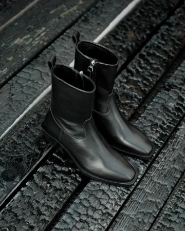 Pair of black leather ankle boots with a zipper closure, positioned on a rustic, charred wooden surface, highlighting a fashion product.