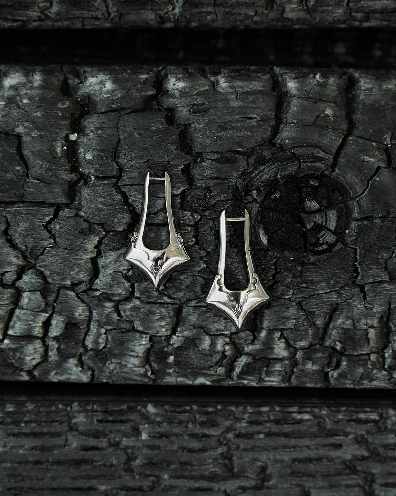 Close-up of silver teardrop earrings on a rough, dark wood surface with visible cracks and textures, jewellery, high-contrast lighting.