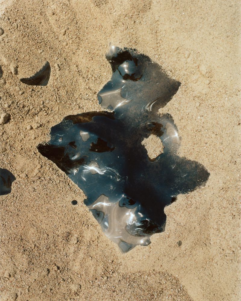 Abstract image featuring a glossy puddle reflecting light on tan sand, with small stones visible around the edges of the puddle.