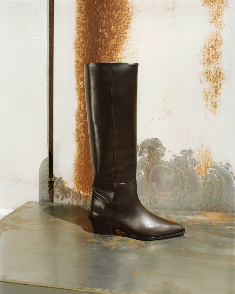 Brown leather knee-high fashion boot on a reflective surface with a mottled, rusted background. Clean, minimalist style.