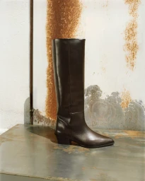 Dark brown leather knee-high boot standing on a distressed metallic surface against a rusted industrial wall.