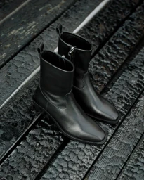 A pair of black leather ankle boots resting on a dark charred wooden surface for the Aeyde Cowboy Capsule project.