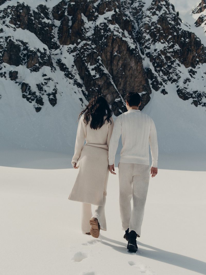 A couple in cream-colored sweaters and trousers walk hand-in-hand across a snow-covered field, with a mountain range rising in the background.