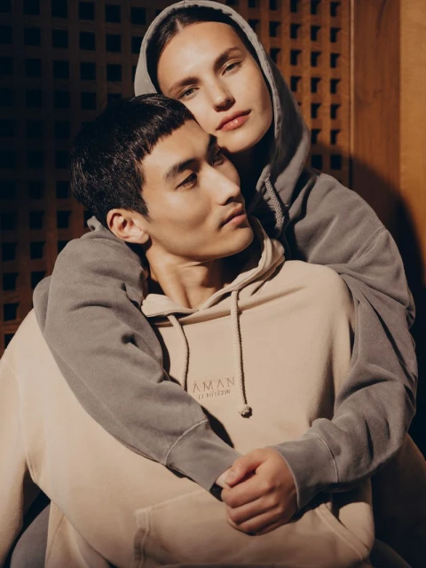 A couple embraces wearing matching hoodies in a studio setting with soft lighting. The female model has her arms wrapped around the male model.