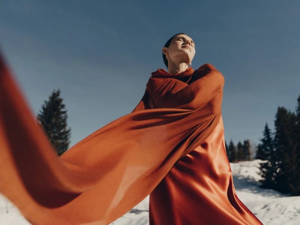 Model wearing a rust-colored silk dress with a flowing matching scarf outdoors in a snowy landscape under a clear sky.