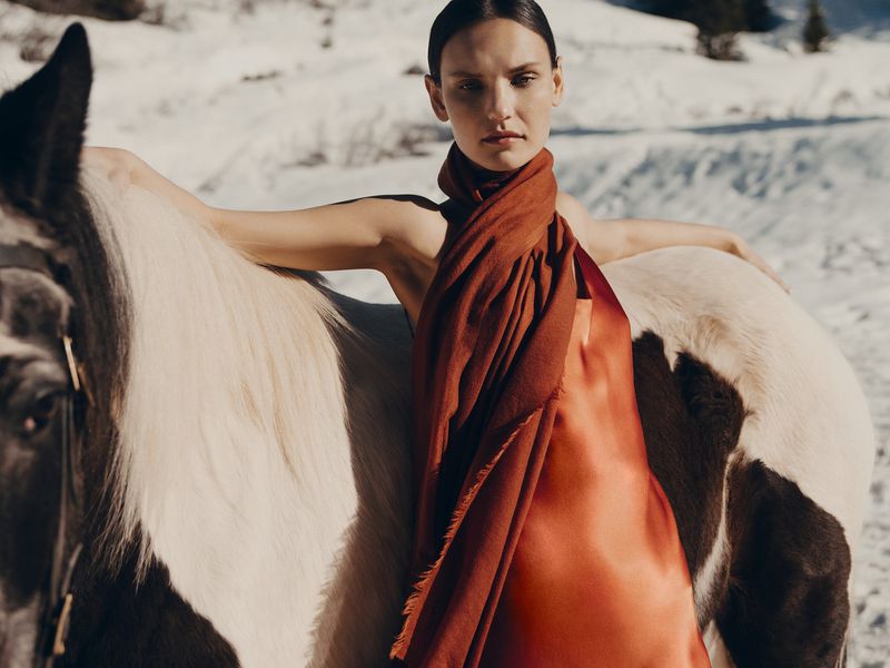 A woman in a red dress and scarf poses with a horse in a snowy field on a sunny day, creating a high-fashion editorial image.