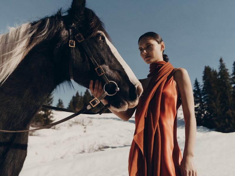 Woman in an orange dress with a scarf posing with a dark horse in a snow-covered landscape with evergreen trees and a blue sky.