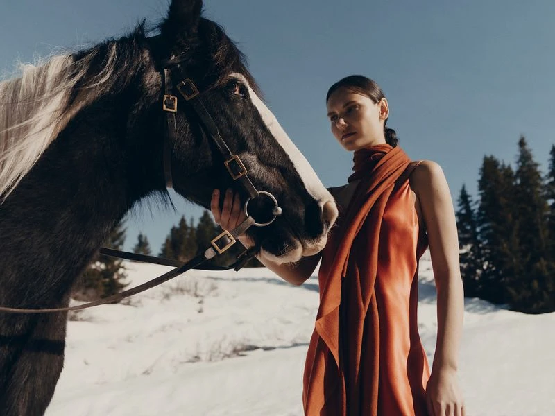 Woman in an orange dress with a scarf posing with a dark horse in a snow-covered landscape with evergreen trees and a blue sky.