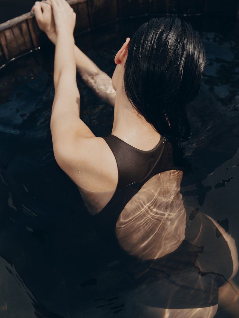 A woman in a black swimsuit is partially submerged in dark water, holding onto the rim of a wooden tub, with light reflections.