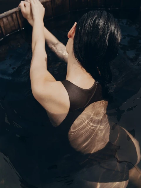 A woman in a black swimsuit is partially submerged in dark water, holding onto the rim of a wooden tub, with light reflections.