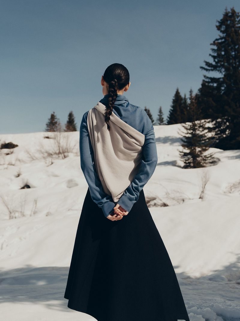 Woman in a black dress and blue top stands in a snowy field with evergreen trees under a blue sky in a fashion shot, with back to camera.